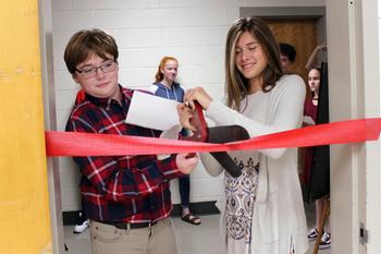a boy and girl cut a red ribbon with a large pair of scissors
