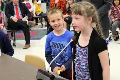 a young girl speaks into a microphone while a little boy looks on