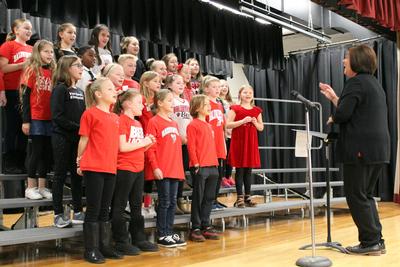 a chorus of young boys and girls dressed in red sing as their teacher directs them