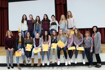 Female volleyball players sit and stand on a stage, holding certificates