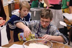 a boy measures cooking oil into a measuring cup as another boy looks over his shoulder