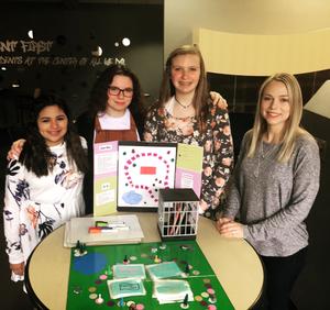 Four females stand behind a table with a board game on it